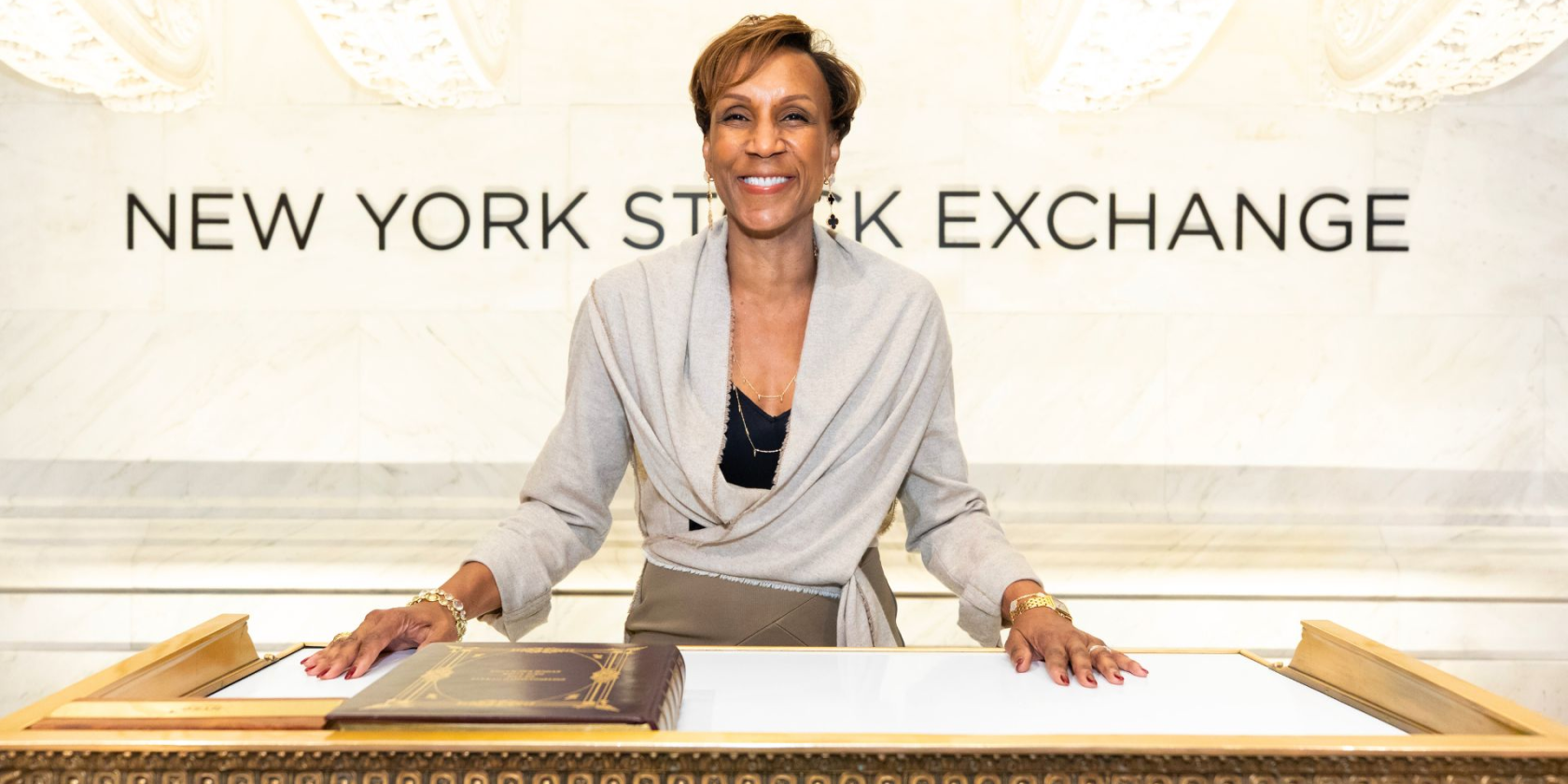 A smiling woman stands behind a podium in front of a sign that reads “NEW YORK STOCK EXCHANGE,” wearing a soft wrap sweater and gold accessories.